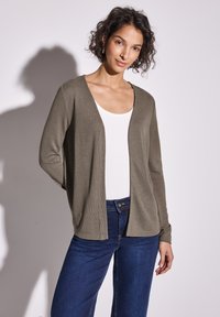 Frau mit kurzen lockigen Haaren trägt eine graue, offene Strickjacke, weißes Oberteil und blaue Jeans, steht vor einem hellen Hintergrund mit Schatten.