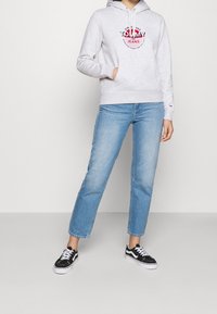 Felpa grigia con logo, tasca frontale, cappuccio regolabile, abbinata a jeans blu chiaro a gamba dritta e sneakers nere. Design semplice e casual.