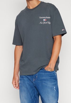 Osoba ubrana w ciemnoszarą koszulkę Tommy Jeans New York City i niebieskie dżinsy, z tatuażem na dłoni i jedną ręką w kieszeni, widoczny tułów.