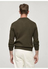 Pull en tricot vert olive avec un col à revers et des poignets côtelés, porté avec un pantalon blanc. Texture lisse, coupe ajustée.