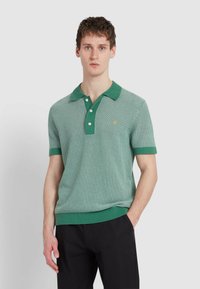 Zielona dzianinowa koszulka polo z kołnierzykiem, teksturowanym wzorem, trzema guzikami i kontrastującymi zielonymi wykończeniami. Noszona na czarnych spodniach.