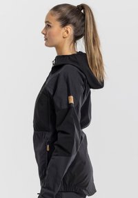 Black lightweight jacket with a hood, made of durable fabric. Features a front pocket, side zip, and leather logo patch on the sleeve.