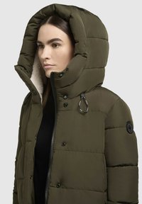 Jachetă pufoasă de culoare verde măslin, cu glugă mare, închidere cu fermoar și accente de hardware negre. Are un căptușeală moale din blană sintetică.