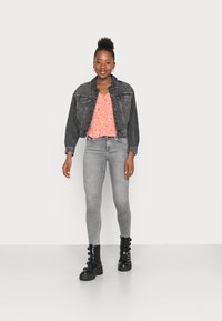 Jaqueta de ganga preta, blusa floral rosa, jeans skinny cinzentos e botas pretas de atar com detalhes em fivela. Fundo simples.