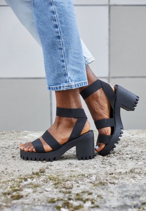 Persona che indossa sandali neri con tacco largo e cinturini spessi, jeans azzurri chiari, in piedi su un terreno dalla texture ruvida.