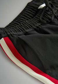 Mustat urheilushortsit, joissa on joustava vyötärö, punavalkoiset sivuraidat ja sileä, teksturoitu kangas.