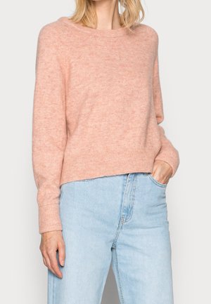 Femme portant un pull à manches longues rose doux et un jean bleu clair taille haute, avec une main dans la poche sur un fond neutre.