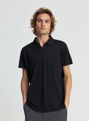 Giovane uomo con capelli biondi e ricci che indossa una semplice polo nera a maniche corte e pantaloni grigi, in piedi con le mani in tasca.