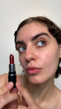 Rouge à lèvres rouge dans un tube noir avec des accents argentés. Le bâton est effilé, et le produit est tenu entre les doigts près du visage.