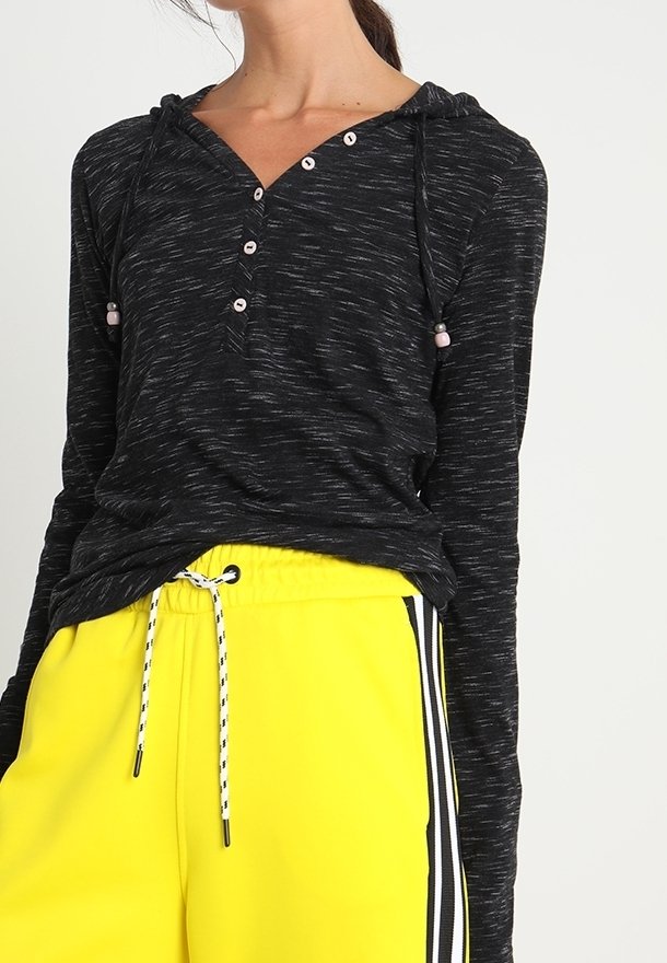 Kvinna som bär en svart långärmad henley-hoodie med knappar och gula dragkedjesvettsbyxor med svarta och vita sidosträck.