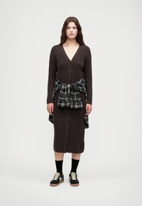 Καφέ ριγέ πλεκτή καλοντή φόρα με v-λαιμόκοψη και κουμπιά στο μπροστινό μέρος, συνδυασμένη με ένα καρό πουκάμισο σε μαύρο και λευκό δεμένο στη μέση και μαύρα αθλητικά παπούτσια.