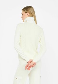 Pulover alb tricotat cu guler înalt, având un model texturat în diamante și mâneci strâmte, purtat peste pantaloni asortați.
