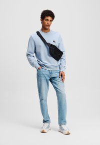 Blauwe sweater, lichtblauwe jeans en een zwarte schoudertas. De sweater heeft een kleine grafische afbeelding. Sneakers zijn wit met blauwe accenten.