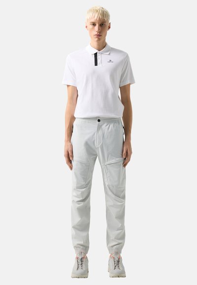 Young person with short blond hair wearing a white polo shirt, light gray cargo pants, and white sneakers, standing facing forward.
