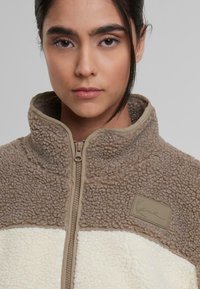 Chaqueta de forro polar en tonos marrón claro y crema. Cuello alto, cremallera completa, tela texturizada con patrón suave. Cuenta con un parche de marca en el pecho.