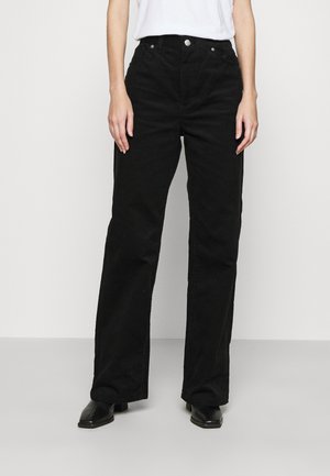 Kobieta ubrana w czarne sztruksowe spodnie z wysokim stanem i szerokimi nogawkami, białą koszulę wpuszczoną do środka oraz czarne buty na kwadratowym nosku, stojąca na jednolitym tle.