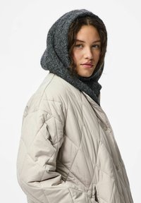 Młoda kobieta w beżowej pikowanej kurtce i grubym szarym dzierganym kapturze, patrząca lekko na bok z neutralnym wyrazem twarzy.