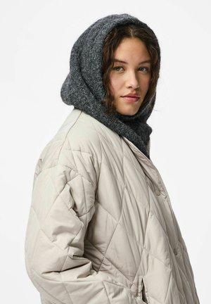 En ung kvinna som bär en vadderad beige jacka och en tjock grå stickad huva, tittar något åt sidan med ett neutralt uttryck.