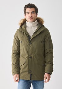 Hombre vistiendo un abrigo parka verde olivo con capucha de piel, suéter de cuello alto beige y jeans azules, de pie contra un fondo claro uniforme.