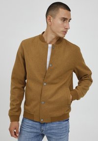 Chaqueta bomber de forro marrón con cuello redondo, botones a presión, bolsillos laterales y puños de canalé, combinada con vaqueros azules.