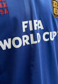 Tissu bleu avec texte blanc en gras « FIFA WORLD CUP », texte partiel rouge à gauche et écusson avec emblème FIFA World Cup noir et or à droite.