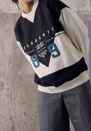 Persona con sudadera Carhartt negra y beige con el número "89" y patrones geométricos, de pie frente a una pared de cemento texturizado.