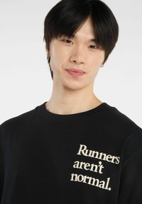 Camiseta de algodón negra con cuello redondo que presenta texto color crema "Los corredores no son normales." en el lado izquierdo. Textura suave.