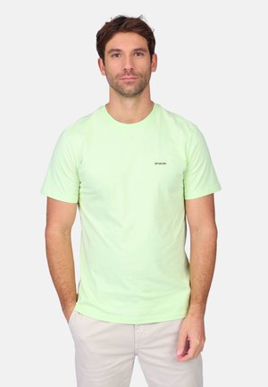 Homme aux cheveux bruns courts portant un t-shirt vert clair à col rond et un pantalon beige clair, debout avec les mains partiellement dans les poches.