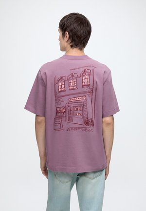 Osoba nosząca fioletowy t-shirt z liniowym rysunkiem fasady księgarni, zawierający okna, drzwi, rower i znak na chodniku.
