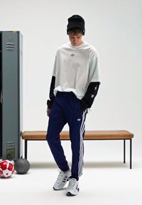 Balta T-krekls ar melnām garām piedurknēm, kurās ir zvaigznes, tumši zili sporta bikses ar baltām svītrām un melni sporta apavi, izvietoti minimālistiskā telpā.