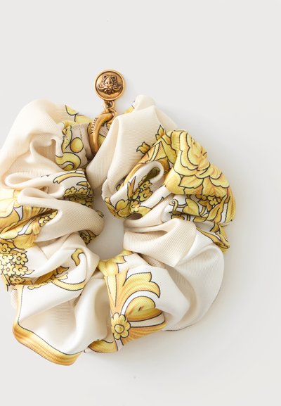Chouchou en soie avec motif floral jaune et baroque et médaillon doré avec logo Versace sur fond blanc.