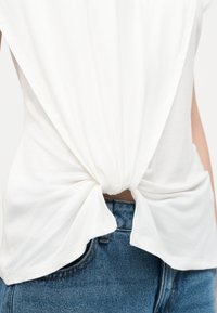 Λευκή μπλούζα με κοντά μανίκια δεμένη κόμπο στη μέση, συνδυασμένη με μπλε τζιν παντελόνι.