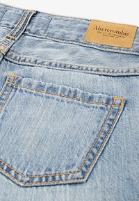 Un material de denim albastru deschis cu cusături vizibile, având un buzunar pe spate și o etichetă din piele bej cu textul "Abercrombie" la talie.