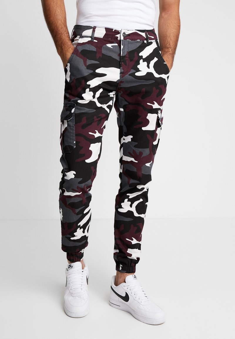 Homme portant un pantalon cargo camouflage noir, blanc, gris et bordeaux avec des bas élastiques et des baskets Nike blanches avec un swoosh noir. Mains dans les poches.