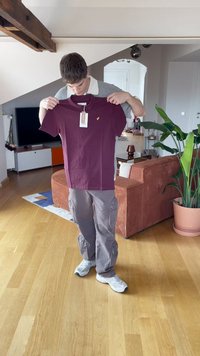 Burgunderfarbenes T-Shirt mit kurzen Ärmeln und gelbem Logo, von einer Person gehalten, mit angehängtem Etikett. Lässiges Design, in einer häuslichen Umgebung präsentiert.