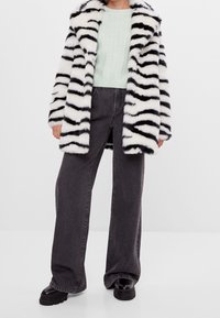 Černobíle pruhovaný umělý kožich s límcem, doplněný světle zeleným texturovaným svetrem a tmavými širokými džínami. Černé robustní boty.