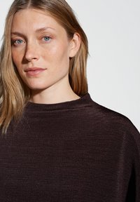 Junge Frau mit heller Haut, blauen Augen und schulterlangem blonden Haar trägt einen dunklen, gerippten Pullover vor einem schlichten weißen Hintergrund.