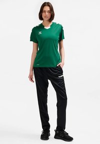 Green short-sleeve sports shirt with a V-neck and logo, paired with black synthetic pants featuring white branding. Athletic footwear completes the outfit.