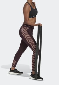 Reggiseno sportivo nero e leggings bordeaux con un motivo a zebra, caratterizzati da una vita alta e un tessuto testurizzato. Persona che tiene delle bande di resistenza.