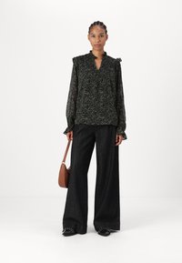 Svart blomstermønstret bluse med volangkrage og ermer, kombinert med vide mørke jeans og en brun crossbody-veske. Svart sko med detaljer.