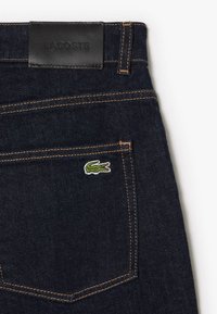 Vaqueros de mezclilla en azul oscuro con un parche de cuero negro etiquetado como "LACOSTE". Presenta un pequeño logo de cocodrilo verde bordado en el bolsillo trasero.