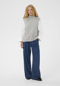 Pull sans manches gris avec col montant, associé à un pantalon en denim bleu à jambes larges. Texture de tissu lisse, détails de couture minimaux.