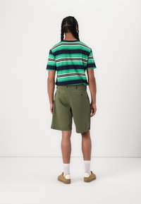 Gestreiftes Kurzarmhemd in Grün, Marineblau und Beige; kombiniert mit olivgrünen Shorts. Braune Schuhe und weiße Socken. Schlichter, lässiger Look.