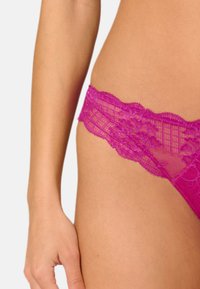 String en dentelle rose vif avec des motifs floraux et des bords festonnés, doté d'une texture transparente et d'une conception de taille lisse.