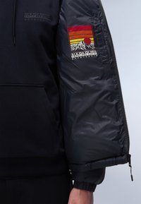 Sort fleece hoodie med en kængurulomme, parret med en polstret sort pufferjakke med et rød- og orange bjergmotiv på ærmet.