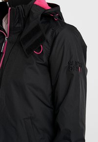 Schwarze wasserdichte Jacke mit pinken Akzenten, ausgestattet mit einem hohen Kragen, einem Reißverschluss vorne und Klettverschlüssen an den Schultern. Markenlogo abgebildet.