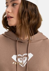 Hnedá mikina s kapucňou so striebornými očkami, so zlatou a bielou srdcovou logom. Mäkkučká tkanina s jemnou textúrou.