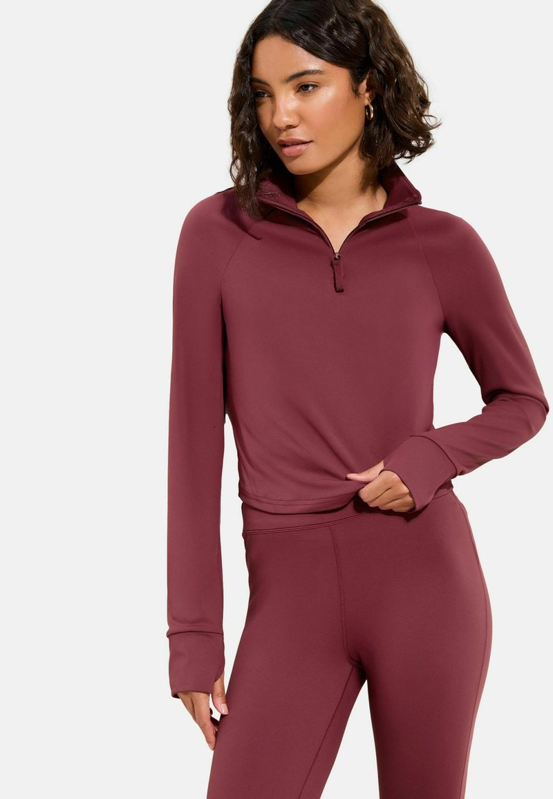 Polaire sportive bordeaux à fermeture éclair quart, manches longues avec ouvertures pour les pouces, associée à des leggings assortis et ajustés. Tissu lisse et extensible.