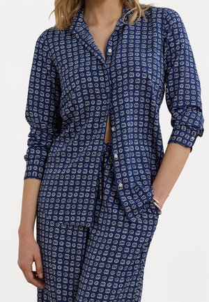 Femme portant une chemise bleue à manches longues boutonnée et un pantalon assorti avec un motif géométrique blanc, une main dans la poche et une sur le côté.