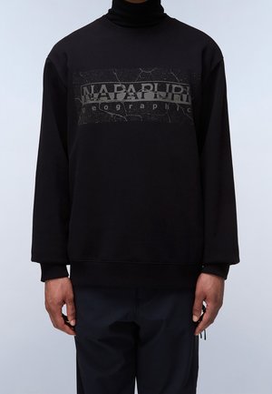 Mies, joka on pukeutunut mustaan huppariin, jossa on "NAPAPIJRI geographic" -teksti ja halkeillut kuvio rinnassa, yhdistettynä mustiin housuihin.